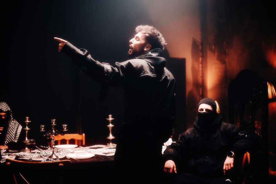 Men in dark attire engaged in intense discussion around a table in dim lighting.
