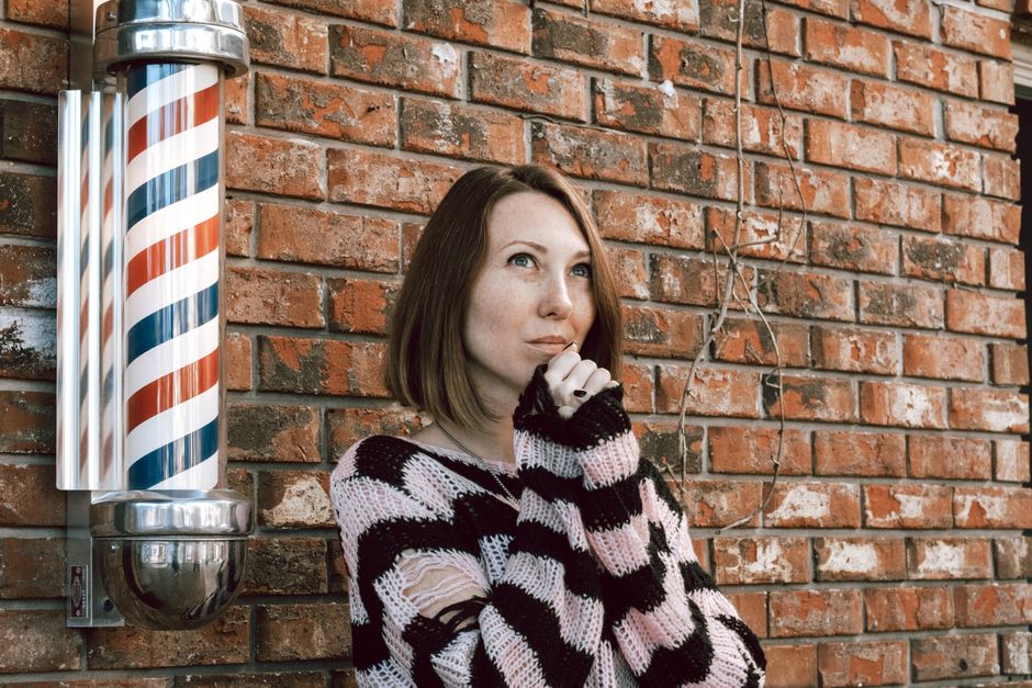 Contemplative woman in a striped sweater stands by a rustic brick wall and barber pole.