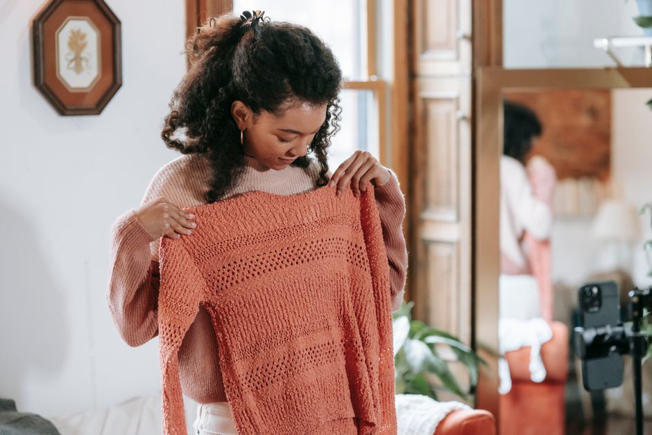 Young African American female with curly hair standing and showing knitted sweater while recording vlog on smartphone in light room