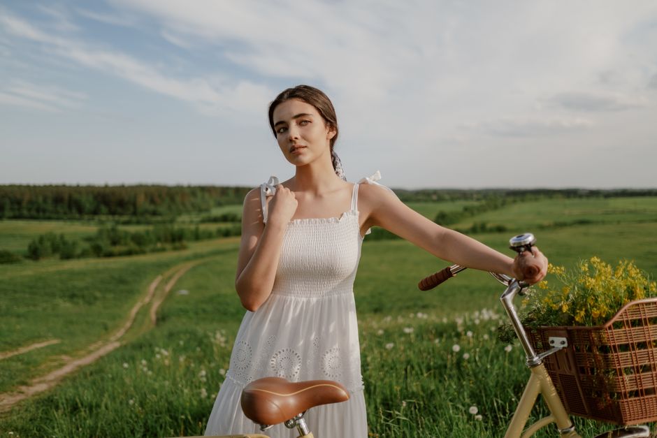 A young woman stands with a bicycle in a lush green meadow under a bright summer sky.