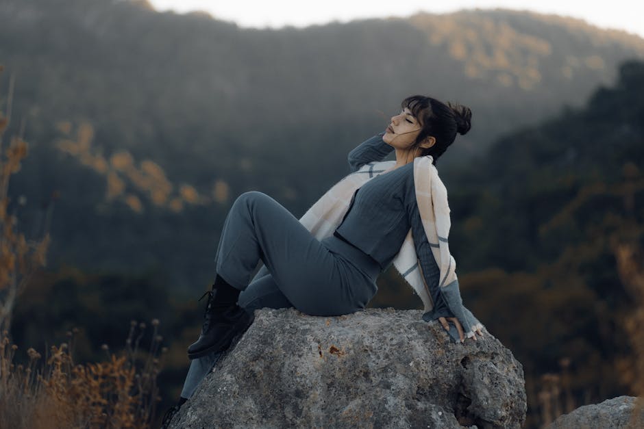 A woman in gray clothing relaxing on a rock amidst a serene mountainous backdrop.