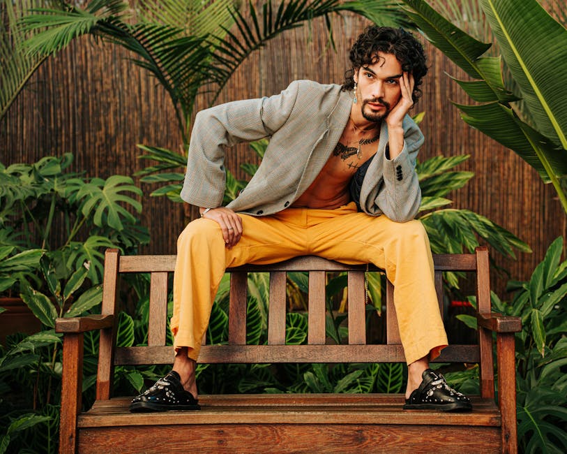 Fashionable man sitting on a wooden bench surrounded by tropical plants, showcasing contemporary style.