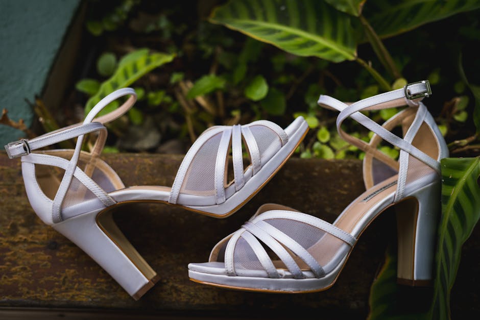 Stylish white strappy high heels resting elegantly on rustic wood amongst leafy greenery.