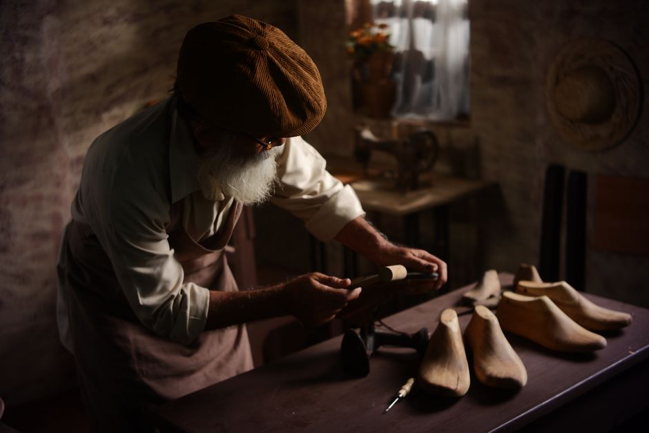 Elderly artisan shaping wooden shoe forms in a vintage workshop setting.