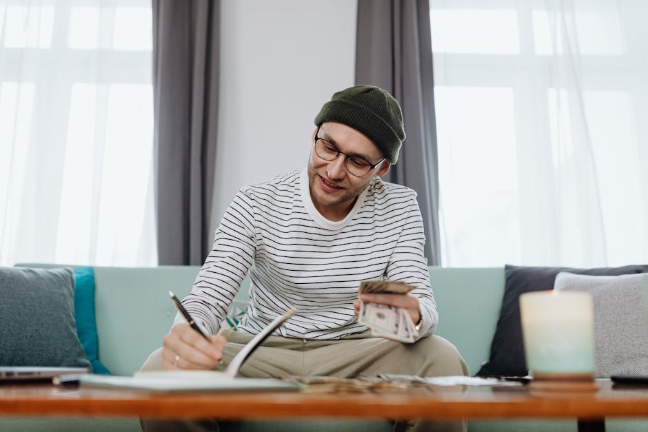 Smiling man in beanie counts cash while writing at home.
