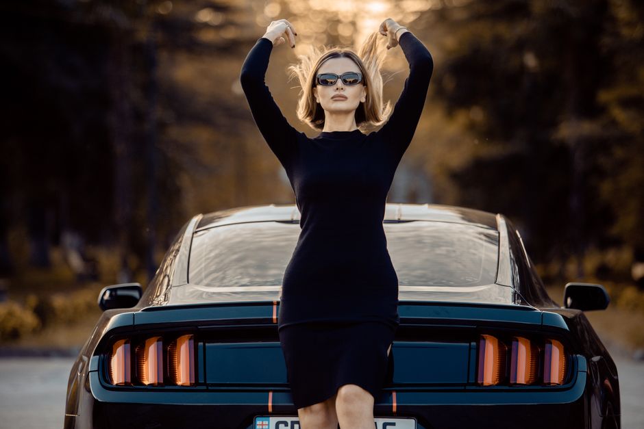 Stylish woman wearing sunglasses poses confidently next to a Ford Mustang in an outdoor setting, embodying elegance and strength.