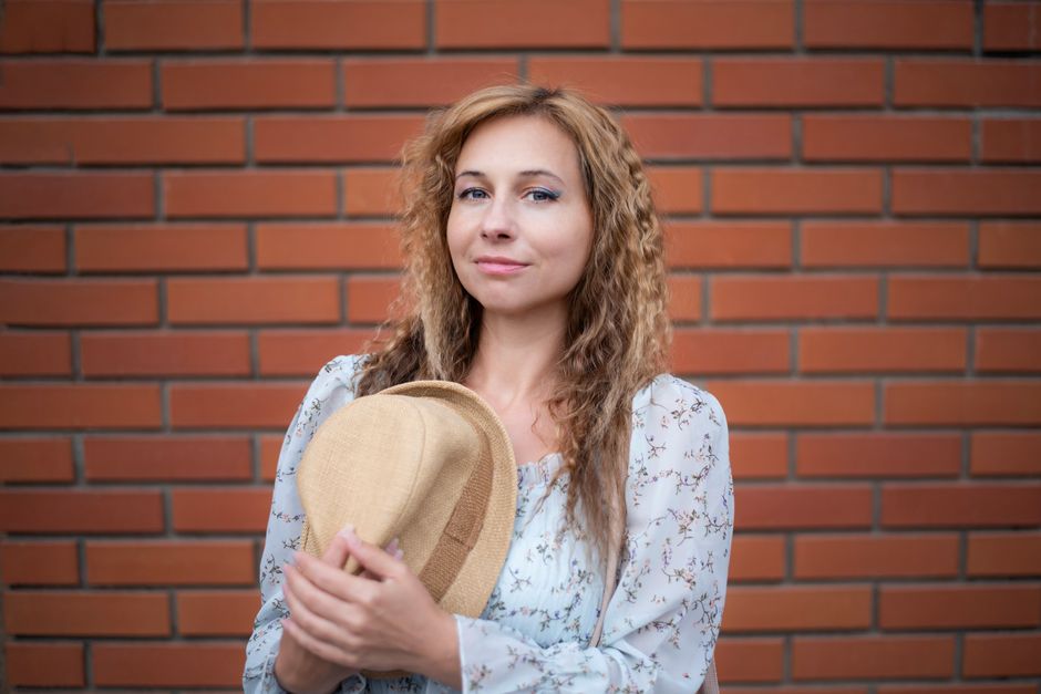 Portrait of a woman holding a hat, standing against a brick wall with a slight smile.