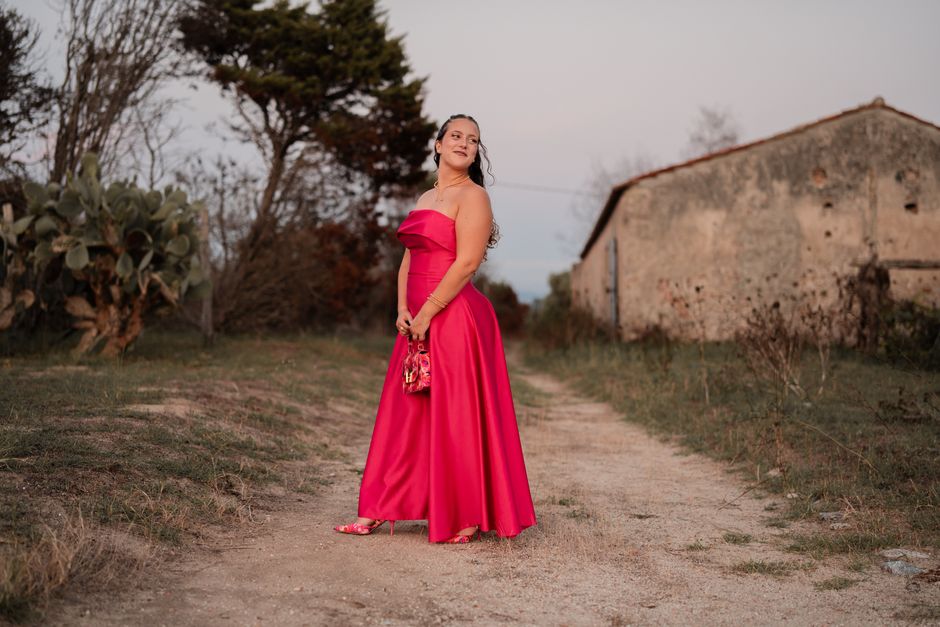 Woman in a vibrant red gown poses gracefully outdoors, blending elegance with rural charm.