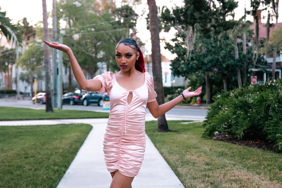 Stylish woman in a pink dress poses confidently on an urban walkway surrounded by greenery.