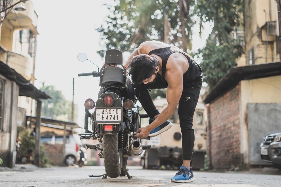 A muscular man adjusts his motorcycle in a sunlit urban alley, showcasing city life and strength.