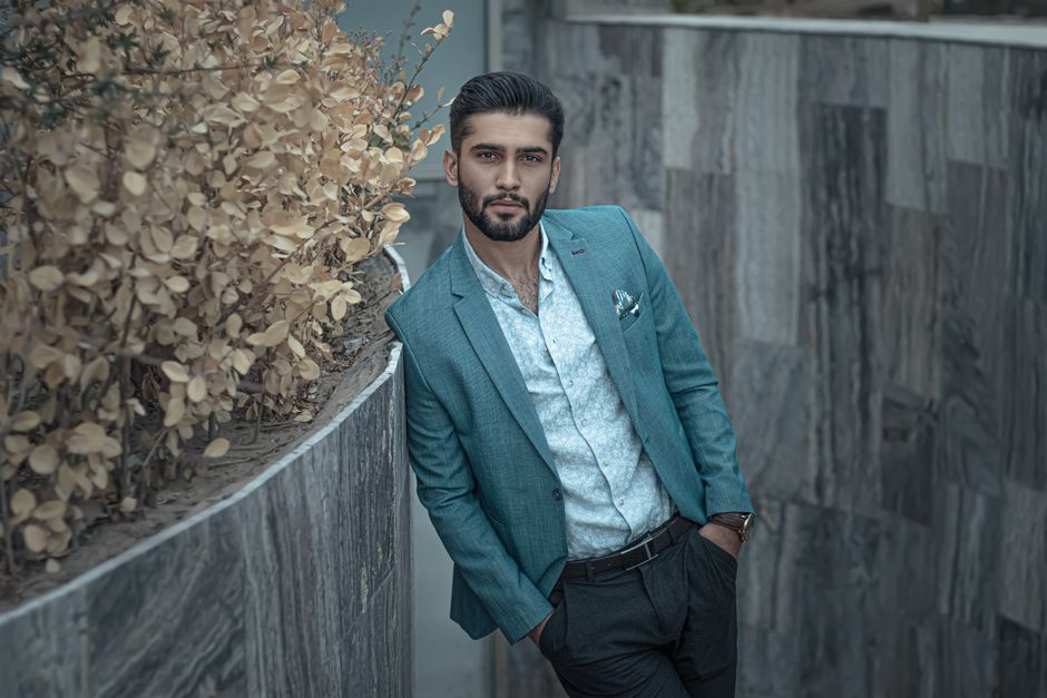 Stylish man in a blue suit poses outdoors against a modern wall in Mashhad, Iran.