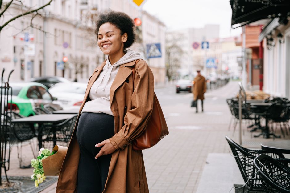 A joyful pregnant woman walking on a city street, holding a bouquet of flowers and smiling.