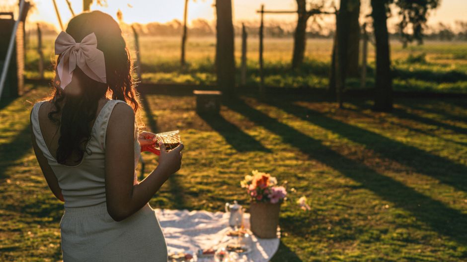 A woman with a pink bow holding a drink at a serene outdoor picnic during sunset.