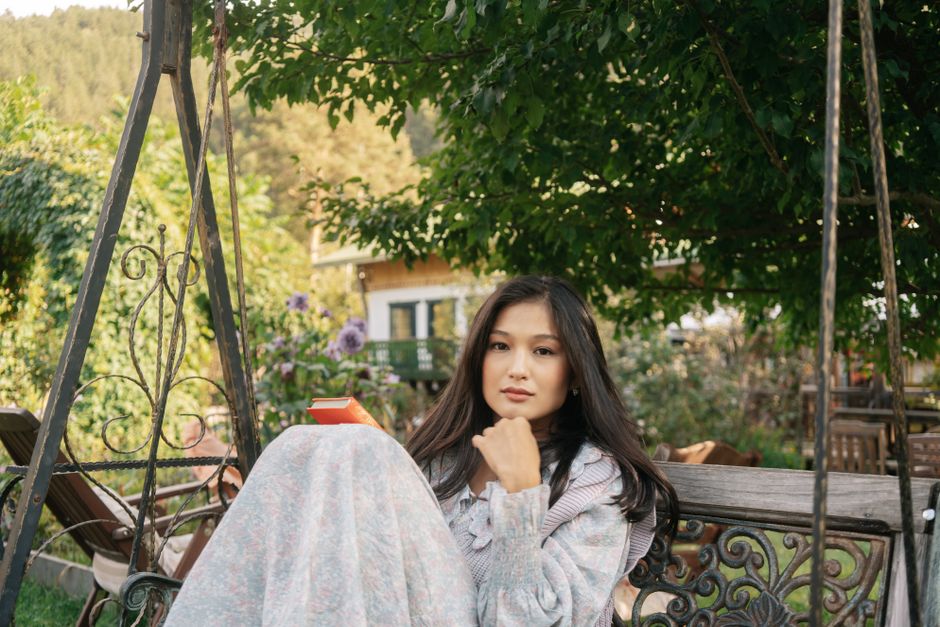 A young Asian woman sits on a swing in a garden, enjoying a book on a peaceful day.