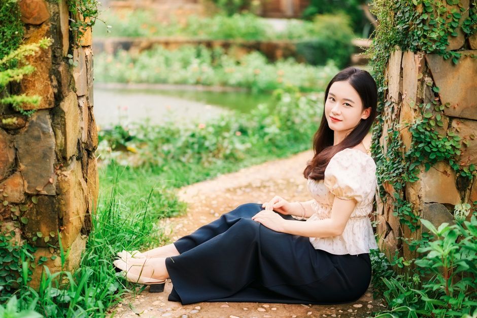 A young woman sits gracefully in a peaceful garden surrounded by lush greenery and stone walls.