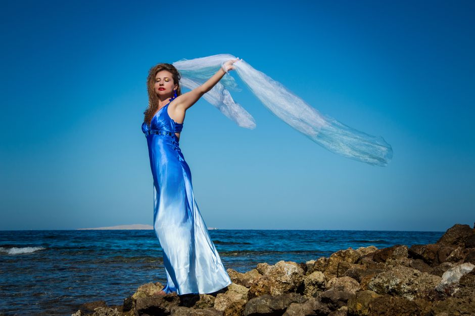 Woman in a flowing blue dress gracefully poses on the rocky seashore.