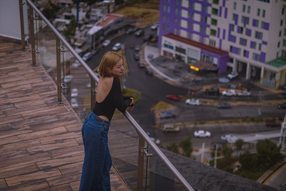 Young woman leans over a rooftop balcony in urban surroundings, city view below.