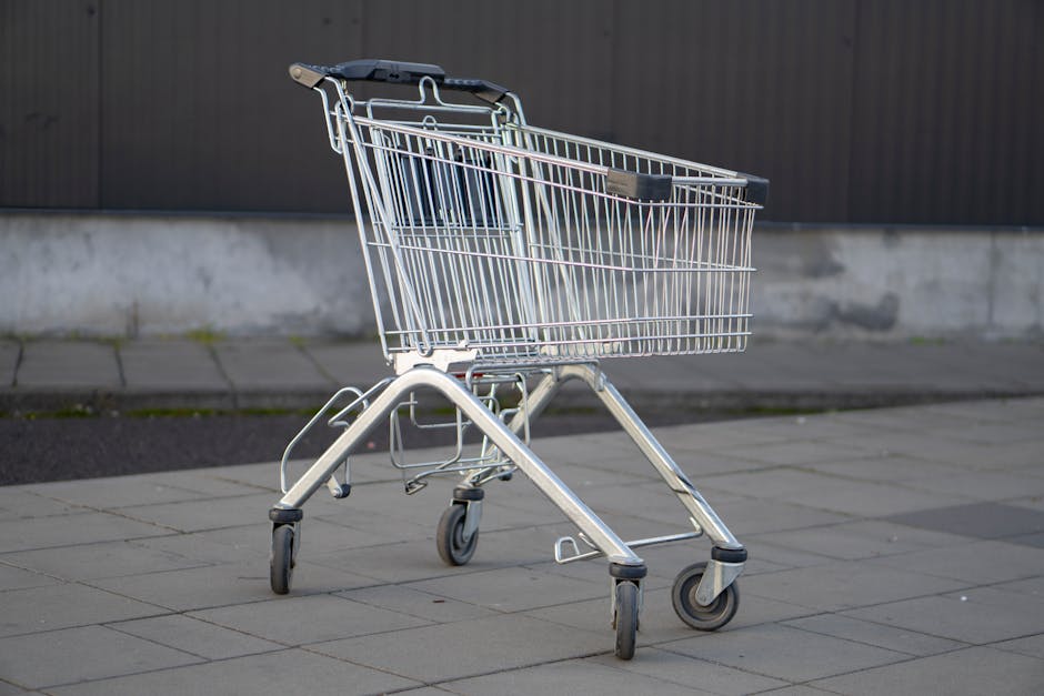 A solitary metal shopping cart stands in an empty outdoor parking lot, capturing the essence of consumerism.