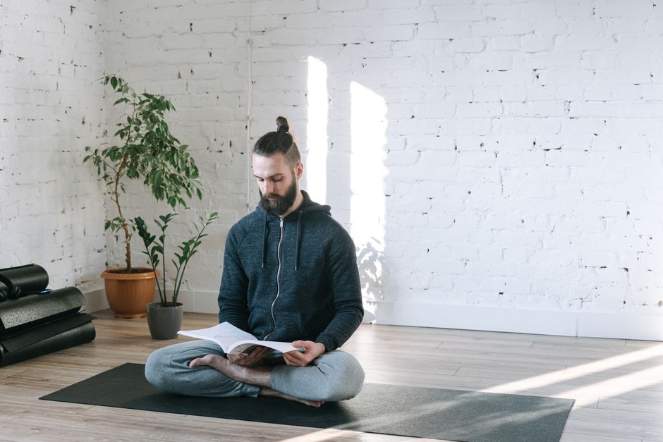 A man reads a book while sitting cross-legged on a yoga mat in a bright, sunlit room.