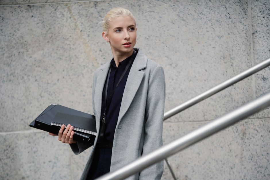 Calm young businesswoman in formal gray coat carrying folders and standing on street while looking away